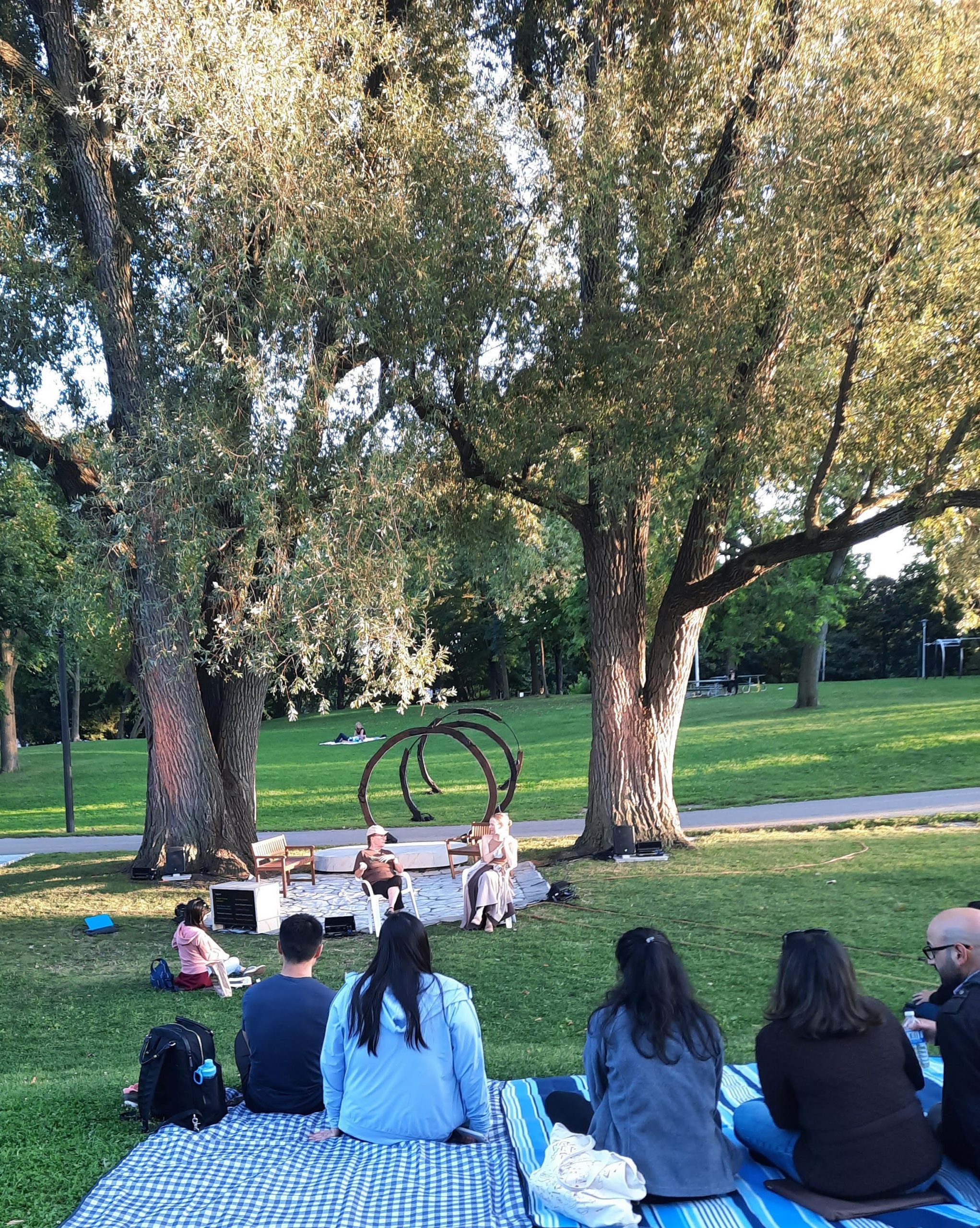 Photo from behind of audience members sitting in a park, facing 2 people talking on the makeshift stage.